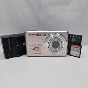 May include: A light pink Casio Exilim digital camera featuring a YouTube capture mode sticker, a silver lens, and the text 12 Mega Pixels. Included are a black battery charger, a Casio NP-20 battery, and a 16GB Evo Plus memory card.