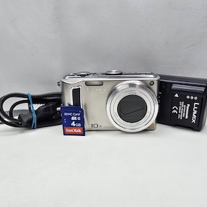 Op de afbeelding: Een zilverkleurige Panasonic Lumix digitale camera met Leica-lens, een zwarte acculader, een zwart netsnoer en een blauwe SanDisk 4GB SDHC-geheugenkaart. De camera heeft een 10x optische zoomlens en de merknaam Lumix op de voorzijde.