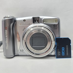 Könnte beinhalten: Eine silberne Canon PowerShot A720 IS Digitalkamera mit 8,0 Megapixel Sensor und 6-fach optischem Zoom. Das Objektiv hat die Spezifikationen 5,8-34,8 mm 1:2,8-4,8. Eine schwarze Samsung 8GB SDHC-Speicherkarte liegt neben der Kamera.
