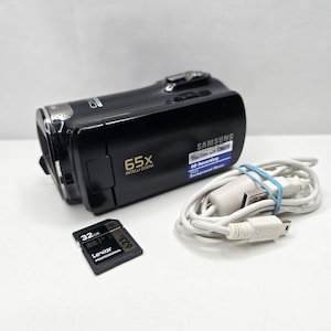Op de afbeelding: Een zwarte Samsung-camcorder met 65x Intelli-zoom, een 32GB Lexar Professional SD-kaart en een witte USB-oplaadkabel. De camcorder is voorzien van een True Color with CMOS HD Recording-label en een Smart Background Music-label op de zijkant.
