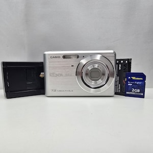 Op de afbeelding: Een zilverkleurige Casio Exilim digitale camera met 7,2 megapixels en een 3x optische zoomlens. De set bevat een zwarte acculader, een zwarte NP-20 lithium-ion accu en een blauwe Traxdata 2GB Secure Digital geheugenkaart.