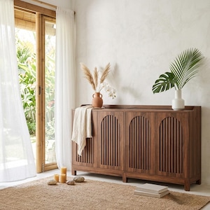 May include: A dark wood cabinet with four doors, each featuring vertical slat detailing. The cabinet is topped with a terracotta vase holding dried grasses and a white vase with a large green leaf. A beige rug and white curtains complete the room.