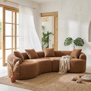 May include: A large, curved brown sofa with several matching throw pillows. The sofa sits on a woven jute rug, and a beige throw blanket is draped over the cushions. A terracotta vase with dried plants is on the rug.