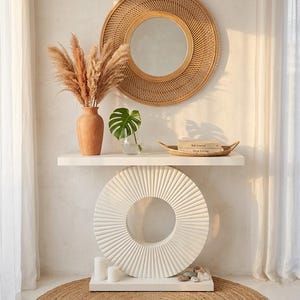 May include: A decorative white console table with a circular, textured design. A round rattan mirror hangs above a white shelf holding a vase of pampas grass, a monstera leaf in a glass, and a woven tray with books. A jute rug is on the floor.