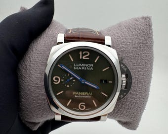 May include: A stainless steel Panerai Luminor Marina automatic watch with a brown leather strap. The watch has a dark green dial with luminous hands and markers. The watch is displayed on a gray pillow.
