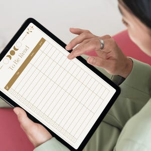 May include: A black tablet displaying a digital "To Be Read" list. The list has a gold header and grid lines, with decorative floral and moon phase elements. The tablet is held by a person wearing a green shirt.