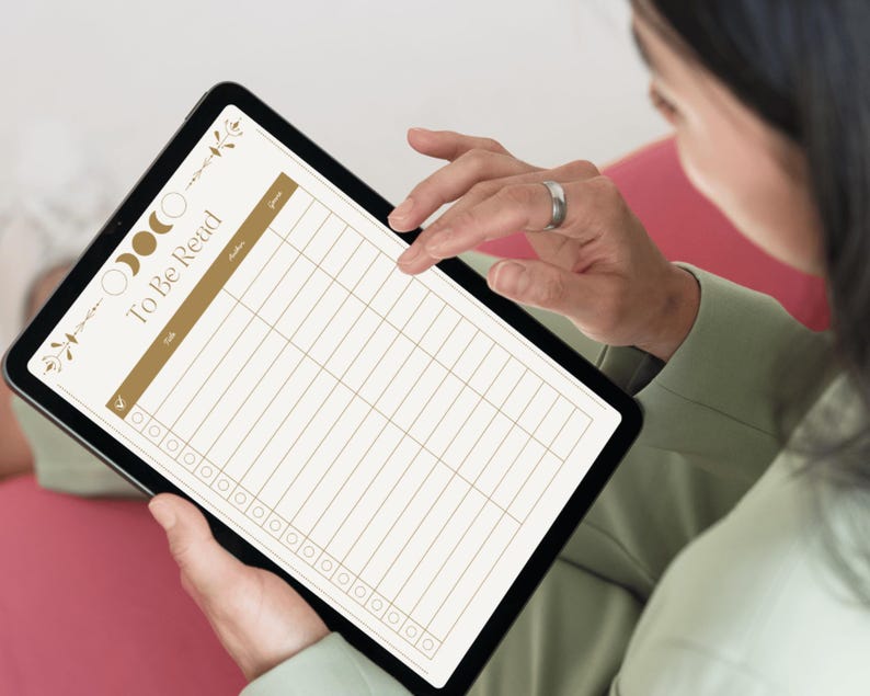 May include: A black tablet displaying a digital "To Be Read" list. The list has a gold header and grid lines, with decorative floral and moon phase elements. The tablet is held by a person wearing a green shirt.