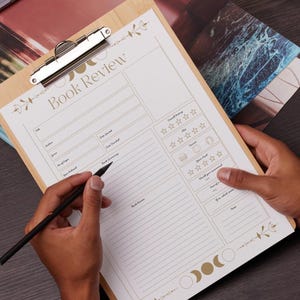 May include: A wooden clipboard with a "Book Review" form printed on it. The form has sections for title, author, and a rating system. A black pen is held over the form, ready to write. The clipboard has a silver clip and decorative floral and moon phase accents.