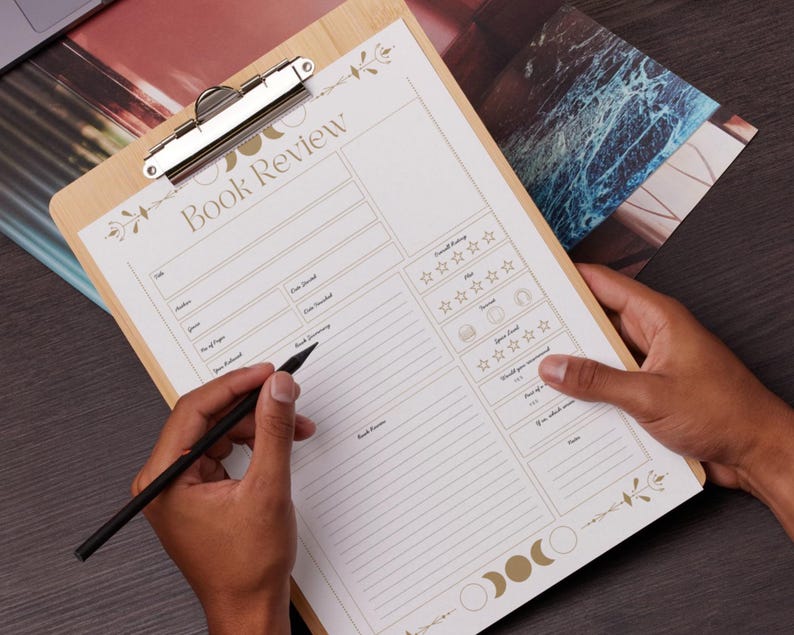 May include: A wooden clipboard with a "Book Review" form printed on it. The form has sections for title, author, and a rating system. A black pen is held over the form, ready to write. The clipboard has a silver clip and decorative floral and moon phase accents.