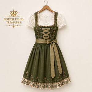 May include: A dark olive green dirndl dress featuring a white lace blouse with short sleeves, a bodice with crisscross ribbon lacing, and a matching olive green apron tied in a bow. The skirt has floral embroidery and a patterned hem border.