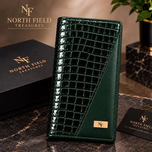 May include: A vertical dark green wallet featuring a glossy crocodile-embossed texture on the left side and a smooth matte finish on the right. A small rectangular gold-toned metal plaque with the letters NF is centred on the lower right side.