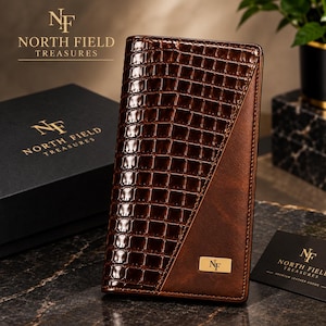 Puede incluir: Una cartera alta y rectangular de color marrón con una textura brillante de piel de cocodrilo en relieve en el lado izquierdo y un acabado marrón mate liso en el derecho. Una pequeña placa rectangular dorada con las letras NF está centrada abajo.
