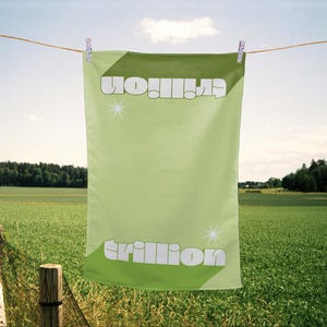 May include: A rectangular light green towel hanging from a clothesline. The towel features the word trillion printed in a bold white sans-serif font at the top and bottom, with small white four-pointed star graphics flanking the text on both sides.