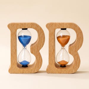 Könnte beinhalten: Zwei hölzerne Sanduhren in Form des Buchstabens B. Eine Sanduhr enthält blauen Sand, die andere orangefarbenen Sand. Die Holzrahmen haben eine hellbraune, natürliche Holzmaserung und die Sanduhren bestehen aus klarem Glas.