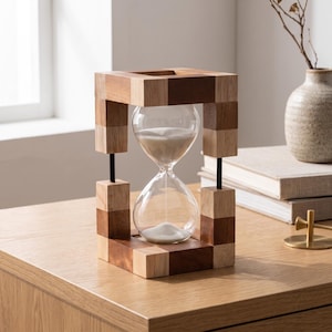 Puede incluir: Un reloj de arena con un bulbo de vidrio transparente lleno de arena blanca, encerrado en un marco rectangular construido con bloques de madera de tonos claros y oscuros, unidos por finas varillas metálicas negras en las esquinas.