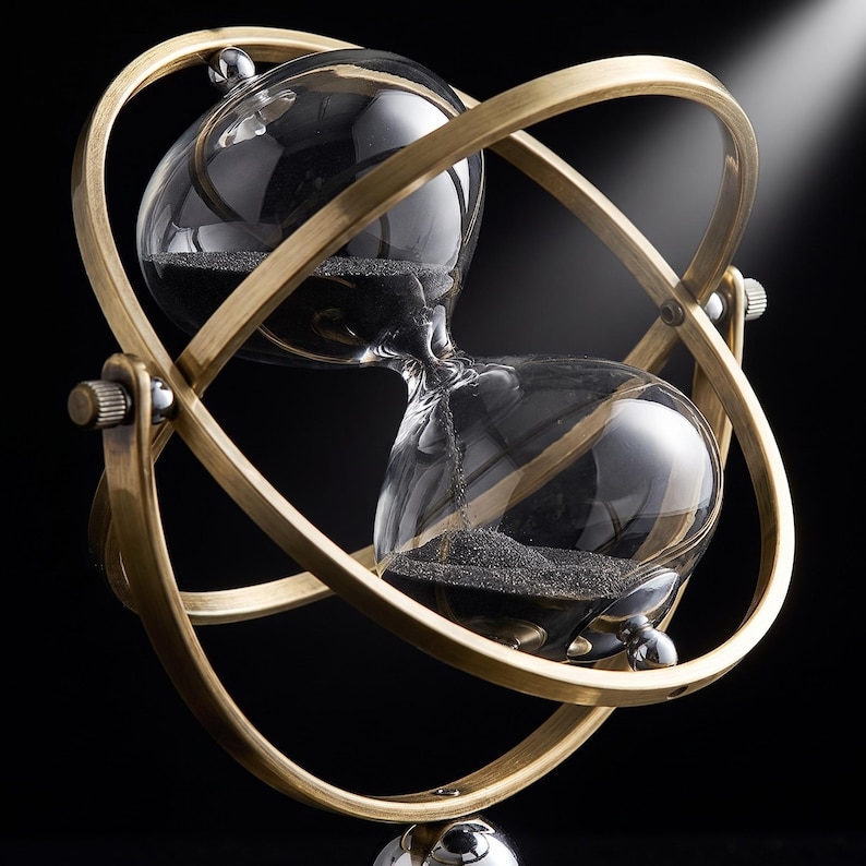 Pu&ograve; includere: Una clessidra con bulbi in vetro trasparente contenenti sabbia nera fine, racchiusa in una cornice metallica rotante color ottone composta da due anelli intersecanti. La struttura presenta piccole manopole metalliche sui lati per la rotazione.