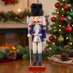 May include: A wooden nutcracker soldier wearing a tall black fuzzy hat, a blue jacket with silver trim and red accents, and blue boots with gold crisscross lacing. The figure holds a silver spear and stands on a rectangular red base.