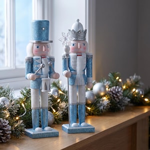 May include: Two decorative nutcracker figures standing on rectangular bases. One nutcracker wears a blue glittery top hat and plays a drum, while the other wears a silver crown and holds a staff topped with a clear snowflake ornament.