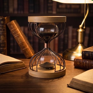 Könnte beinhalten: Eine Sanduhr mit einem klaren Glaskolben, der mit dunklem Sand gefüllt ist und von einem kupferfarbenen Metalldrahtrahmen umschlossen wird. Die Struktur wird durch kreisförmige, helle Holzsockel an der Ober- und Unterseite gestützt.