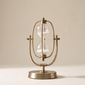 Puede incluir: Un reloj de arena giratorio con un marco de metal de latón cepillado y un bulbo de vidrio transparente que contiene arena blanca. El reloj de arena está montado sobre una base circular y sostenido por una estructura tipo cardán de 360 grados.