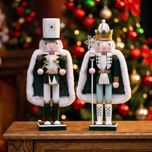 May include: Two nutcracker figures wearing dark green coats with white faux fur trim. The left figure wears a white top hat with a snowflake and holds a drum. The right figure wears a gold crown and holds a staff topped with a snowflake ornament.