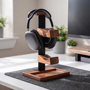 May include: A vertical headphone stand featuring a black metal central pillar, two curved wooden rests for headphones, and a rectangular wooden base with a recessed tray. A pair of dark grey over-ear headphones rests on the top wooden support.