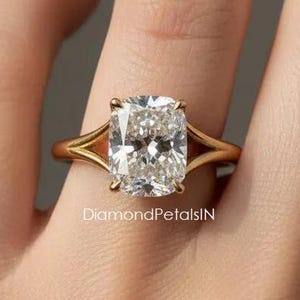 May include: A gold ring featuring a large, rectangular cushion-cut clear gemstone held by four prongs on a split shank band. The text DiamondPetalsIN is visible on the band. The design showcases a polished metal finish and a faceted stone.