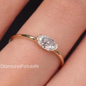 Könnte beinhalten: Ein dünner Goldring mit einem einzelnen horizontalen, oval geschliffenen, klaren Edelstein in einer Zargenfassung. Das Band ist glatt und poliert, wobei der Text DiamondPetalsIN in kleiner weißer Schrift nahe der Edelsteinfassung zu sehen ist.