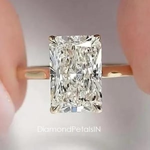 Könnte beinhalten: Ein Ring mit einem Diamanten im Radiant-Schliff, bestehend aus einem großen, rechteckigen, klaren Edelstein auf einem dünnen, polierten Gelbgoldring. Der Text DiamondPetalsIN ist in kleinen, weißen, serifenlosen Buchstaben unter der Fassung gedruckt.