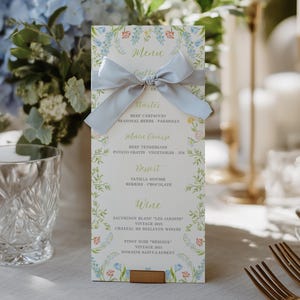 May include: A vertical rectangular menu card featuring a light blue satin ribbon tied in a bow across the top. The card has a white background with a delicate floral border in blue, green, and red, and lists courses including beef carpaccio.
