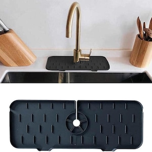 May include: A black silicone sink mat with a rectangular design and vertical slots. The mat is placed under a gold-colored faucet in a kitchen sink. A wooden knife block and utensil holder are visible.