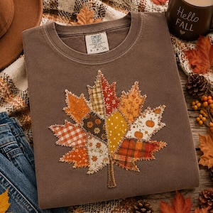 May include: A brown crewneck sweatshirt featuring a large, central maple leaf appliqué made from various fabric patches in orange, yellow, and white patterns, including gingham, floral, and pumpkin prints, with frayed edges and visible stitching.