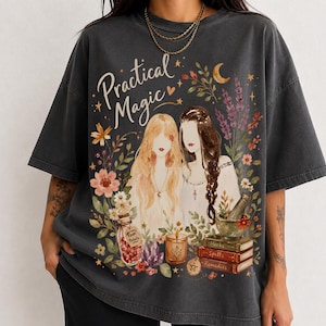 May include: A charcoal gray t-shirt featuring a graphic print of two figures with long hair, surrounded by flowers, a crescent moon, stars, a bottle labeled Rose Petals, a candle, a mortar and pestle, and books labeled Herbs, Spells, and Remedies.