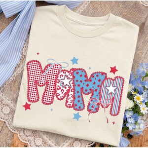 May include: A cream-colored t-shirt featuring the word MAMA in large, stylized letters. The letters are decorated with red polka dots, red and white leopard print, blue star patterns, and red and white vertical stripes, accented with white stars and bows.