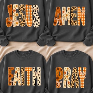 Könnte beinhalten: Ein dunkelgraues Sweatshirt mit Rundhalsausschnitt und vier verschiedenen Grafikdesigns mit den Wörtern JESUS, AMEN, FAITH und PRAY. Jeder Buchstabe ist mit einer Mischung aus orangefarbenen Blumen, Leopardenmuster, orangefarbenem Karomuster und schwarzen Punkten sowie kleinen Schleifen gefüllt.