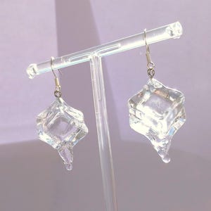 Op de afbeelding: Transparante, geometrische oorhangers met een uniek, getextureerd ontwerp. De oorbellen hangen aan zilverkleurige haken en worden gepresenteerd op een transparante standaard tegen een zachte paarse achtergrond. De oorbellen hebben een driedimensionale, diamantachtige vorm.