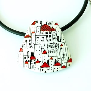 Könnte beinhalten: Halskette mit einem weißen Keramik-Anhänger und einer schwarzen Schnur. Der Anhänger zeigt ein handgemaltes Design einer Stadtlandschaft mit weißen Gebäuden und roten Dächern.