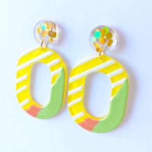 Op de afbeelding: Een paar hangende oorbellen met ronde knopjes gevuld met goudkleurige, iriserende confetti. Hieraan hangen grote, gele, ovale hangers versierd met diagonale witte strepen, een lichtgroen gedeelte en een klein koraalkleurig accent.