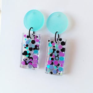 May include: A pair of earrings with a clear, rectangular pendant filled with colourful confetti in black, purple, teal, and blue. The pendants hang from a light blue circular stud with a black hook. The earrings are handmade.