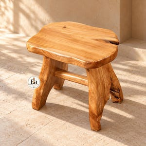 May include: A light brown wooden stool with a natural live edge top and four sturdy legs connected by a horizontal support beam. A circular logo featuring the letters EH and a small torch icon is visible on the front left leg of the stool.