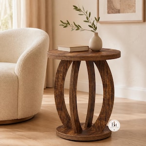 May include: A round wooden side table with a dark brown wood grain finish, featuring a circular top and a base supported by three curved, arch-shaped legs connected to a circular bottom platform. A small beige book and a vase with greenery sit on top.
