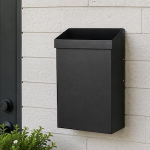 May include: A matte black wall-mounted mailbox featuring a rectangular vertical body and an open-top slanted design. The mailbox has a clean, solid construction with visible side screws and a smooth finish, mounted securely on a light-colored brick wall.