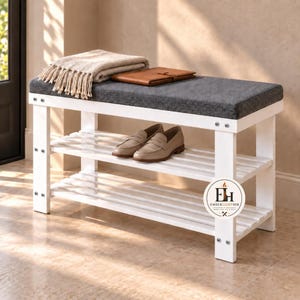 May include: A white wooden shoe bench with two slatted shelves and a dark gray upholstered seat. A beige fringed throw blanket and a brown leather notebook rest on top. A circular logo with the text EmberLightHub is visible on the front leg.