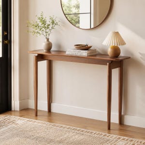 May include: A long, narrow wooden console table with four tapered legs. On top sits a light-colored ceramic vase with green foliage, a small wooden bowl with keys, two stacked books titled This is Home and Live Beautiful, and a small lamp.