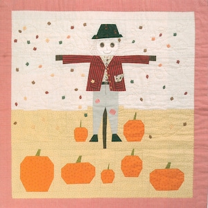 May include: A patchwork quilt featuring a scarecrow standing in a field of pumpkins. The scarecrow is wearing a red and white striped jacket, grey trousers, and a black hat. The pumpkins are orange and have green stalks.