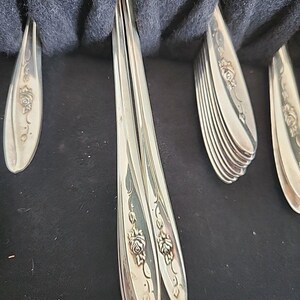 May include: A set of silver-toned metal flatware handles arranged in a dark storage case. Each handle features a raised floral rose motif with leaves and stems embossed along the center. The metallic surfaces are polished and reflective.