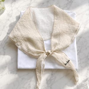 May include: A beige fabric collar with a white lace trim edge, featuring a tie-front design and the embroidered text "Mrs. Garcia" on one of the pointed ends. The collar is layered over a plain white garment, showcasing a textured weave.
