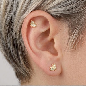May include: A pair of gold stud earrings shaped like butterflies, each featuring a crescent moon design on one wing and small, clear, round-cut stones embedded along the wings. One earring is positioned in the earlobe and one in the upper cartilage.