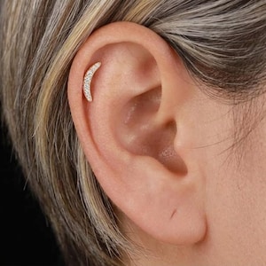 May include: A gold-toned crescent moon earring featuring a row of small, clear, sparkling stones set along the curved surface. The piece is positioned in the upper cartilage area of the ear, showcasing a delicate and curved design.