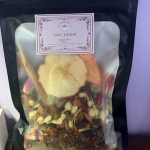May include: A black resealable pouch containing a dried botanical blend with a label reading LOVE POTION SIMMER POT SPELL. The contents include a large dried apple slice, cinnamon sticks, small white flower buds, and various dried herbs and petals.
